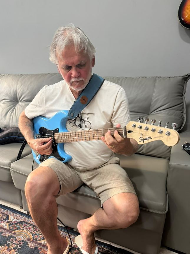 Aluno de cabelo branco tocando uma guitarra azul no sofá da sala